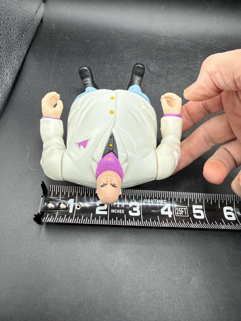 May include: A vintage action figure of a character in a white coat, purple tie, and blue pants. The figure is approximately 3 inches long. The figure has black shoes and is lying on its back.