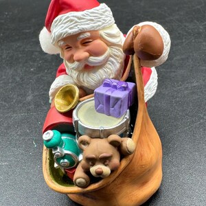 Könnte beinhalten: Eine Weihnachtsmann-Figur mit rotem Hut und Mantel, weißem Bart und rosigen Wangen. Er hält einen Sack voller Spielzeug, darunter ein Teddybär, eine Trommel und ein verpacktes lila Geschenk. Oben befindet sich eine goldfarbene Öse.