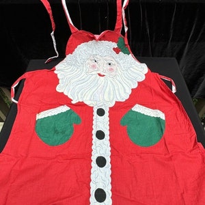 May include: Red Christmas apron with a white Santa Claus face, white beard, and black buttons. The apron has green mitten pockets on each side.