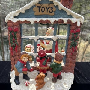 May include: A ceramic Christmas toy shop with a red roof and a sign that says "TOYS". Santa Claus is inside the shop, and three children are looking in the window. There is a dog in front of the shop.