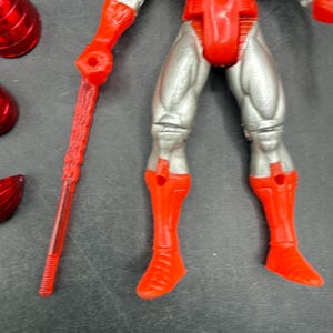 Puede incluir: Una figura de acci&oacute;n roja y plateada con un bast&oacute;n rojo. La figura tiene botas, guantes y armadura pectoral rojos, con piernas y brazos plateados. Varios accesorios rojos est&aacute;n esparcidos alrededor de la figura.