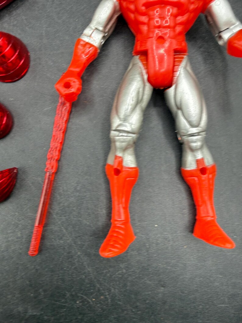 Puede incluir: Una figura de acci&oacute;n roja y plateada con un bast&oacute;n rojo. La figura tiene botas, guantes y armadura pectoral rojos, con piernas y brazos plateados. Varios accesorios rojos est&aacute;n esparcidos alrededor de la figura.