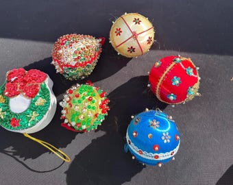 May include: A collection of colorful Christmas ornaments. The ornaments include a wreath, a red and green beaded ornament, a yellow ornament with red accents, a red ornament with blue floral accents, a green and yellow beaded ornament, and a blue ornament with white and blue accents.