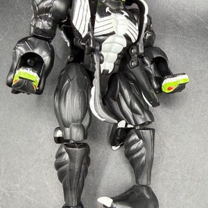 May include: A black and white action figure with a green and red head. The figure has a muscular build with articulated joints. The figure's chest has a white design. The figure's feet have white claws.