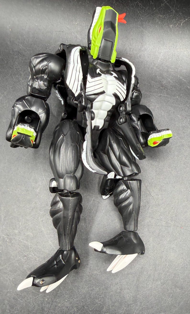 May include: A black and white action figure with a green and red head. The figure has a muscular build with articulated joints. The figure's chest has a white design. The figure's feet have white claws.