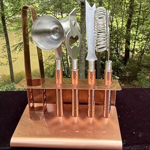 May include: A bar tool set featuring a copper-colored stand holding a jigger, a bottle opener, a knife, and a strainer. Each tool has a textured copper handle. The set includes a pair of tall copper tongs positioned on the left side of the stand.