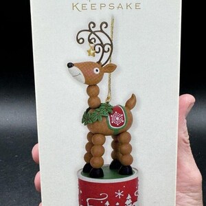 以下が含まれることがあります： ホールマーク・キープセイクのクリスマスオーナメント。茶色と緑のトナカイが、赤と白の雪の結晶のデザインで飾られています。トナカイは、赤と白の台座の上に立っており、冬の風景が描かれています。オーナメントのタイトルは「Wiggle-Wobble Reindeer」です。