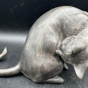 Puede incluir: Una figurita de cerámica gris de un gato enroscado en posición de dormir. El gato tiene una superficie texturizada y una cara detallada.