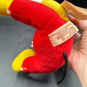 May include: A vintage Mickey Mouse plush toy, primarily red with yellow accents on the ears and feet. The toy has a black tail and a tag with text that reads "THE WALT DISNEY CO." and "MADE IN CHINA".