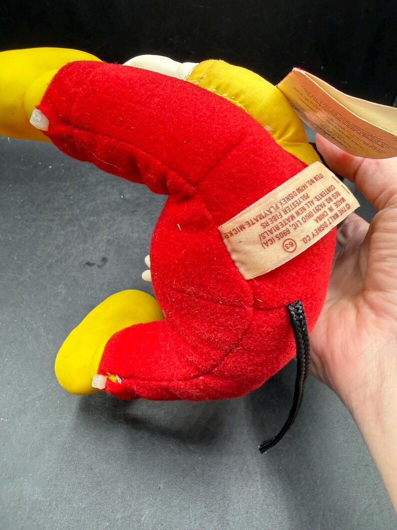 May include: A vintage Mickey Mouse plush toy, primarily red with yellow accents on the ears and feet. The toy has a black tail and a tag with text that reads "THE WALT DISNEY CO." and "MADE IN CHINA".