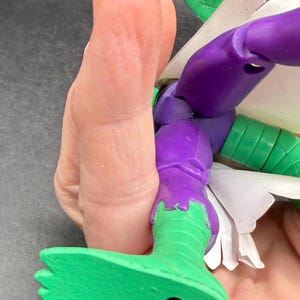 K&ouml;nnte beinhalten: Eine teilweise sichtbare Actionfigur mit gr&uuml;n und lila bemalten Details. Der Fu&szlig; und das Bein der Figur sind gr&uuml;n und gehen am Knie in Lila &uuml;ber. Eine Hand h&auml;lt die Figur, wobei im Hintergrund eine gr&uuml;ne Hand zu sehen ist.