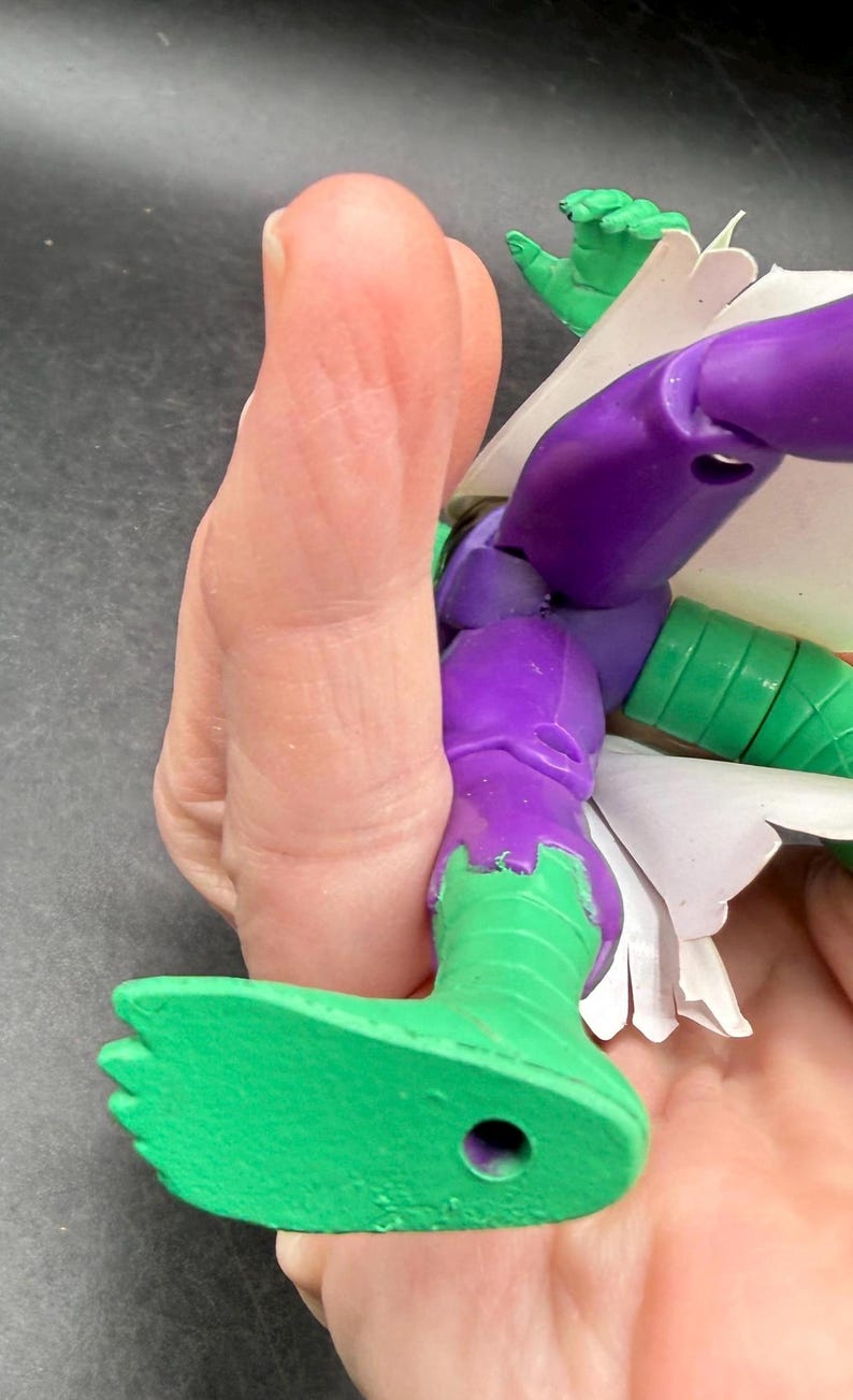 K&ouml;nnte beinhalten: Eine teilweise sichtbare Actionfigur mit gr&uuml;n und lila bemalten Details. Der Fu&szlig; und das Bein der Figur sind gr&uuml;n und gehen am Knie in Lila &uuml;ber. Eine Hand h&auml;lt die Figur, wobei im Hintergrund eine gr&uuml;ne Hand zu sehen ist.