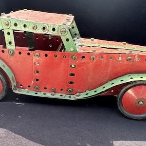 May include: A red and green toy car made of metal with a detailed construction. The car has a rounded body and a long bonnet. The car is made of metal and has a lot of rivets and screws.