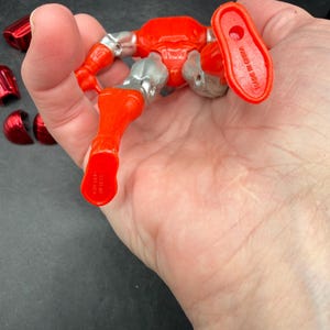Puede incluir: Una figura de acci&oacute;n roja y plateada con articulaciones, sostenida en una mano. La figura tiene una apariencia met&aacute;lica con armadura roja y detalles plateados. Varias piezas rojas separadas son visibles cerca. Las palabras "MADE IN CHINA" est&aacute;n impresas en el pie de la figura.