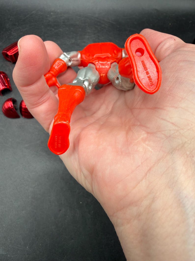 Puede incluir: Una figura de acci&oacute;n roja y plateada con articulaciones, sostenida en una mano. La figura tiene una apariencia met&aacute;lica con armadura roja y detalles plateados. Varias piezas rojas separadas son visibles cerca. Las palabras "MADE IN CHINA" est&aacute;n impresas en el pie de la figura.
