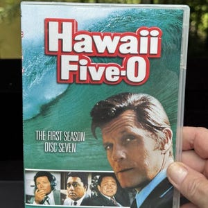 May include: DVD case for the television show "Hawaii Five-0". The case features the show's title in red and white, a wave graphic, and a photo of a man. Text on the case reads "The First Season Disc Seven."