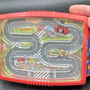 May include: A vintage red plastic toy with a race track design. The toy features a clear plastic top, a winding road, and small toy cars. The words "LEFT TURN" and "RIGHT TURN" are visible on the sides.