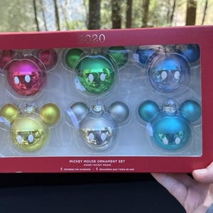 Può includere: Un set di sei decorazioni natalizie di Topolino in una scatola rossa. Le decorazioni sono in vari colori, tra cui rosa, verde, blu, giallo e argento. La scatola è etichettata con "2020" e "Mickey Mouse Ornament Set".