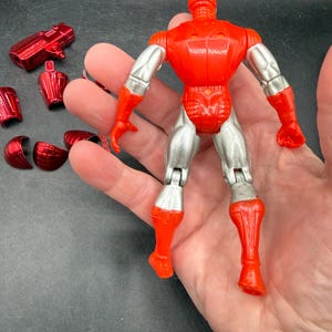 Puede incluir: Una figura de acci&oacute;n roja y plateada con piezas de armadura desmontables. La figura tiene un torso, cabeza y piernas rojos, con brazos y piernas inferiores plateados. Varias piezas de armadura rojas est&aacute;n dispersas.