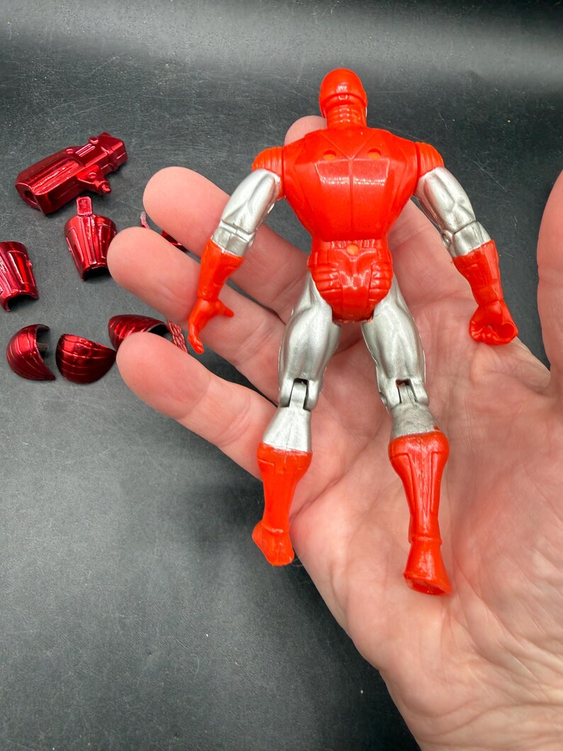 Puede incluir: Una figura de acci&oacute;n roja y plateada con piezas de armadura desmontables. La figura tiene un torso, cabeza y piernas rojos, con brazos y piernas inferiores plateados. Varias piezas de armadura rojas est&aacute;n dispersas.