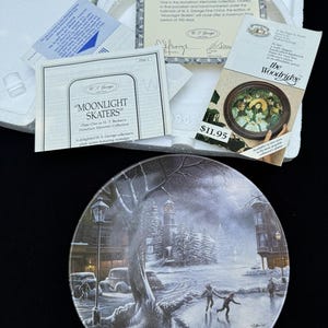 Peut inclure: Une assiette de collection en porcelaine intitulée Moonlight Skaters représentant une scène de ville hivernale avec des patineurs, accompagnée d'un certificat d'authenticité, d'une fiche descriptive et d'un dépliant publicitaire pour un cadre.