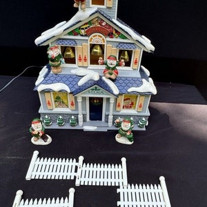 Puede incluir: Una casa de Navidad de varios pisos, azul y blanca, con un techo cubierto de nieve. La casa tiene un letrero que dice "S. Claus" y "Taller". Hay pequeños duendes en el techo y frente a la casa. La casa está rodeada por una valla blanca de madera.