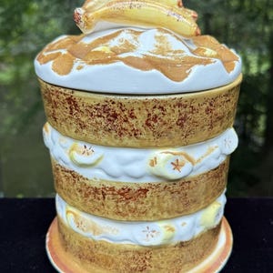 May include: A cylindrical ceramic cookie jar shaped like a layered banana cake. The exterior features textured brown cake layers with white frosting bands decorated with sculpted banana slices. The lid is topped with two sculpted yellow bananas.