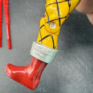 以下が含まれることがあります： 黒い格子線が入った黄色のフィギュアの脚のクローズアップ。フィギュアは赤いブーツと「&copy;1994 MARVEL CHINA」とプリントされたシルバーのバンドが付いています。2つの赤いプラスチックアクセサリーが背景にあります。