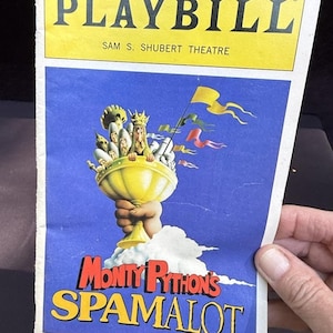 Puede incluir: Un programa de mano de Monty Python's Spamalot, con una pancarta amarilla con "PLAYBILL" y el teatro Sam S. Schubert. La portada tiene un fondo azul con un cáliz dorado sostenido por una mano y el título del espectáculo.