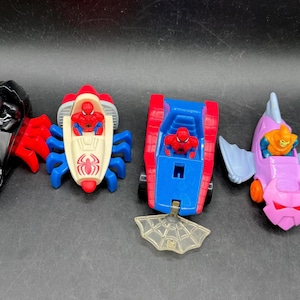 May include: Four colourful toy vehicles featuring Spider-Man and other characters. Includes a black and red vehicle, a red and blue spider-themed car, a red and blue tank-like vehicle, and a purple and blue fish-shaped vehicle.