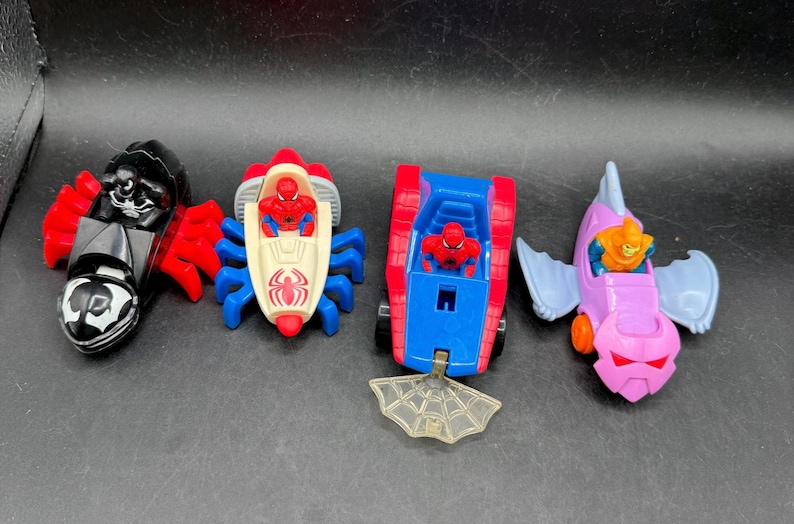 May include: Four colourful toy vehicles featuring Spider-Man and other characters. Includes a black and red vehicle, a red and blue spider-themed car, a red and blue tank-like vehicle, and a purple and blue fish-shaped vehicle.