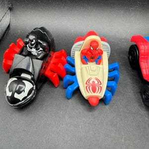 May include: Three toy vehicles featuring Marvel characters. The first is black with red accents and a Venom figure. The second is cream, red, and blue with a Spider-Man figure. The third is red and blue with black wheels.