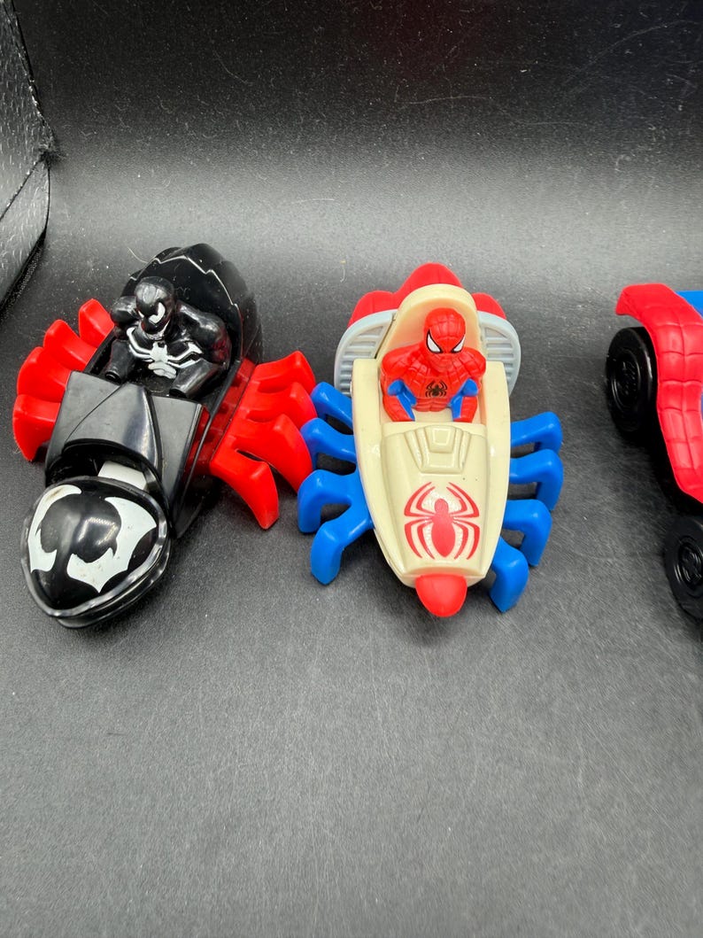 May include: Three toy vehicles featuring Marvel characters. The first is black with red accents and a Venom figure. The second is cream, red, and blue with a Spider-Man figure. The third is red and blue with black wheels.