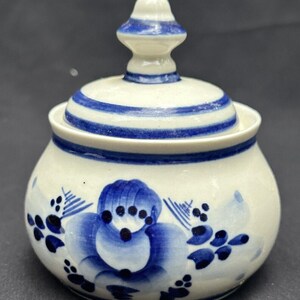 May include: A white ceramic sugar bowl with a blue and white floral design and a lid with a knob. The bowl has a round shape and a blue and white striped band around the top.
