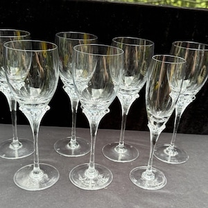 May include: A set of seven clear crystal wine glasses. Each glass has a long stem and a decorative design at the base of the bowl. The glasses are arranged on a dark surface, showcasing their elegant design.