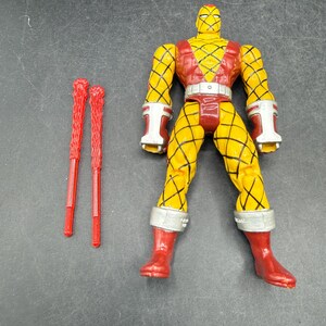 以下が含まれることがあります： 黄色と赤のアクションフィギュア。ダイヤモンド模様、白いアクセント、赤いブーツが特徴です。2本の赤い質感のあるバトンがフィギュアの左側に配置されています。コレクション用の玩具です。