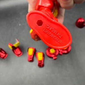 May include: A red and yellow Iron Man action figure, with several detached pieces. The figure's foot is visible, with the text "&copy;1995 MARVEL TOY BIZ, INC." printed on it. The figure is held by a person.