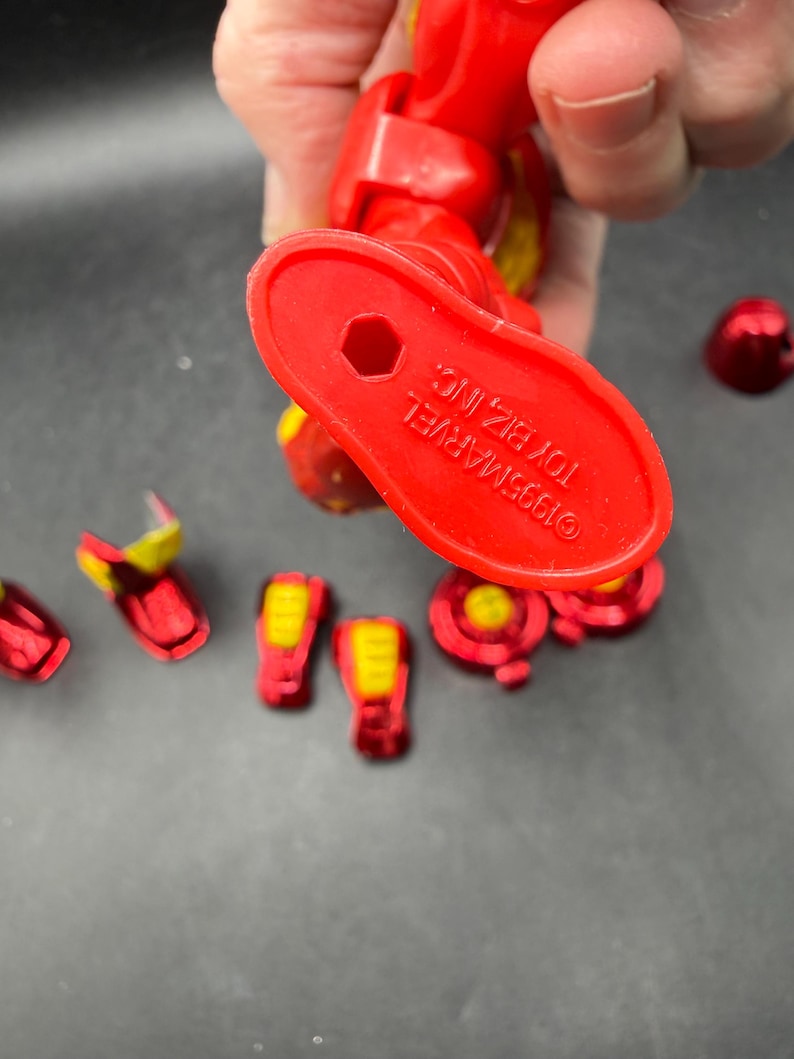 May include: A red and yellow Iron Man action figure, with several detached pieces. The figure's foot is visible, with the text "&copy;1995 MARVEL TOY BIZ, INC." printed on it. The figure is held by a person.