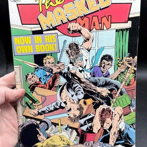 May include: A comic book titled "The Masked Man" with a dynamic action scene on the cover. The cover features a red circle with yellow text, and the phrase "Now in His Own Book!" is also visible. The comic is marked with a price of $1.75 in the USA.