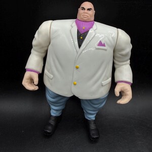 May include: A Kingpin action figure. The figure is wearing a white suit with a purple ascot and light blue pants. The figure has black shoes and a bald head. The suit has yellow buttons and a purple pocket square.