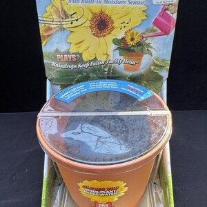 May include: A brown plastic flower pot with a clear plastic lid. The pot has a label that says "Alerts You When Plant Needs Water!" and "Try Me!" The pot is sitting on a green and white background. The pot is part of a singing flower kit that plays "Raindrops Keep Fallin' On My Head".