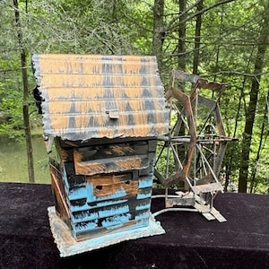 May include: A rustic, decorative water mill sculpture. The miniature building is painted black, blue, and brown with a weathered appearance. A metal water wheel is attached to the side. The sculpture is set against a natural background.