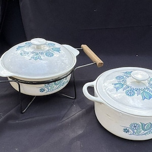 Puede incluir: Dos platos para hornear de cerámica blanca con diseños florales azules y verdes. El plato más grande está en un soporte de metal negro con asas de madera.