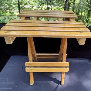 Könnte beinhalten: Kleiner, zusammenklappbarer Holztisch mit natürlicher Oberfläche. Der Tisch hat eine Lattenoberfläche und ein unteres Regal, das von abgewinkelten Beinen getragen wird. Der Tisch ist für den Außenbereich konzipiert und misst ca. 61 cm in der Länge.