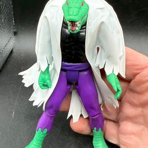 K&ouml;nnte beinhalten: Eine gr&uuml;ne, echsenartige Actionfigur mit wei&szlig;em Laborkittel, schwarzem Hemd und lila Hose. Die Figur hat einen detaillierten Reptilienkopf mit roten Akzenten und ist mit ausgestreckten Armen posiert.