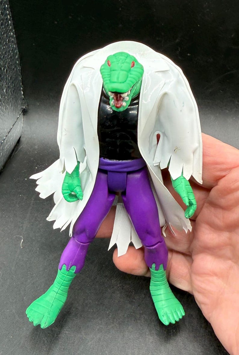 K&ouml;nnte beinhalten: Eine gr&uuml;ne, echsenartige Actionfigur mit wei&szlig;em Laborkittel, schwarzem Hemd und lila Hose. Die Figur hat einen detaillierten Reptilienkopf mit roten Akzenten und ist mit ausgestreckten Armen posiert.