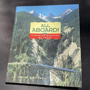 May include: A hardcover book titled "ALL ABOARD! The Canadian Rockies by Train" featuring a scenic mountain landscape with a train crossing a bridge. The cover has a yellow and red banner with the title and author's name.