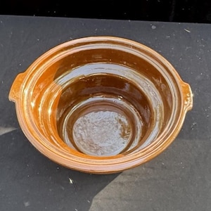 Puede incluir: Un cuenco redondo de cerámica esmaltada con un acabado brillante en color ámbar marrón. El cuenco presenta una amplia abertura circular con un borde escalonado y dos pequeñas asas laterales integradas que sobresalen del borde superior.