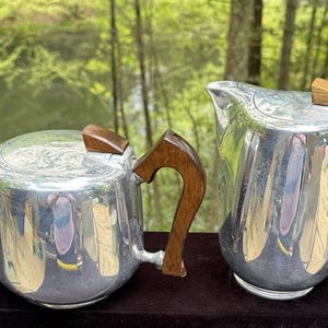 May include: A silver tea set with wooden handles and lid accents. The set includes a teapot and a creamer, both with a reflective surface. The wooden handles are curved and add a warm contrast to the silver.