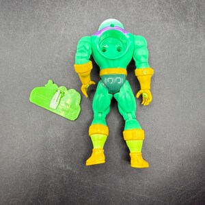 Op de afbeelding: Groene actiefiguur met gele accenten, waaronder handschoenen, laarzen en een riem. De figuur heeft een paarse helm en een groen accessoire. Vintage verzamelobject.